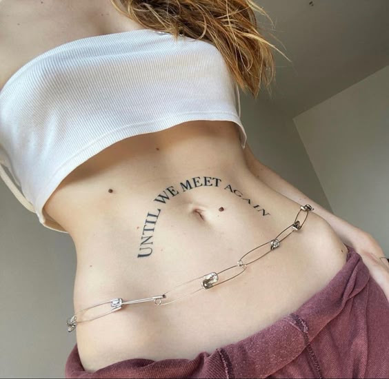 Tattooed abdomen with Until We Meet Again, wire belt, white crop top; unique body art and fashion statement.