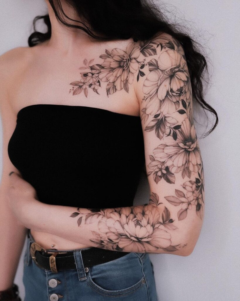 Woman with intricate floral tattoo sleeve in black ink wearing a black tube top and high-waisted jeans.