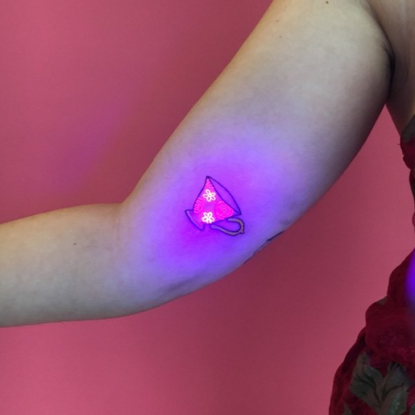 A black light tattoo of a teacup, with the details glowing brightly under UV light, showcasing the subtle daytime appearance and vibrant blacklight effect.