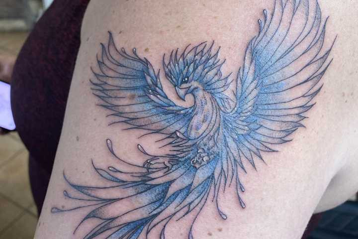 A serene blue phoenix tattoo on a woman's upper arm, utilizing cool blue tones to symbolize spirituality, enlightenment, inner peace, and a unique expression of strength through body art.