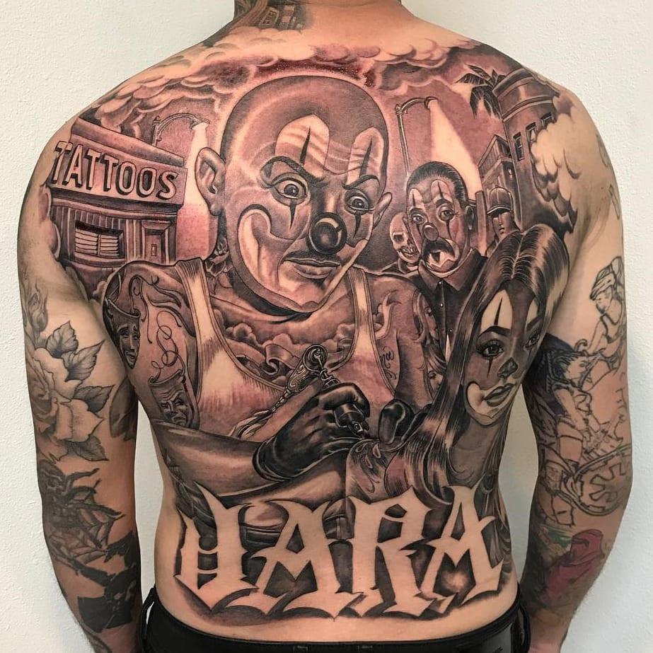 Tattoo by Mister Cartoon. Backpiece Chicano tattoo by Mister Cartoon featuring payasos, a street scene, and lettering in black and grey, demonstrating the large-scale artistry and storytelling in Mister Cartoon's Chicano back tattoos.