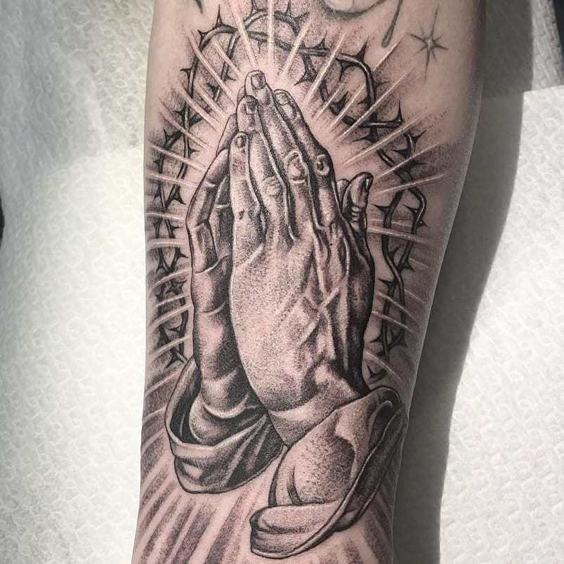 Tattoo by Em Scott. Serene Chicano tattoo by Em Scott depicting praying hands with a crown of thorns and light rays in black and grey, showcasing religious iconography and fine line work in Em Scott's Chicano tattoo style.