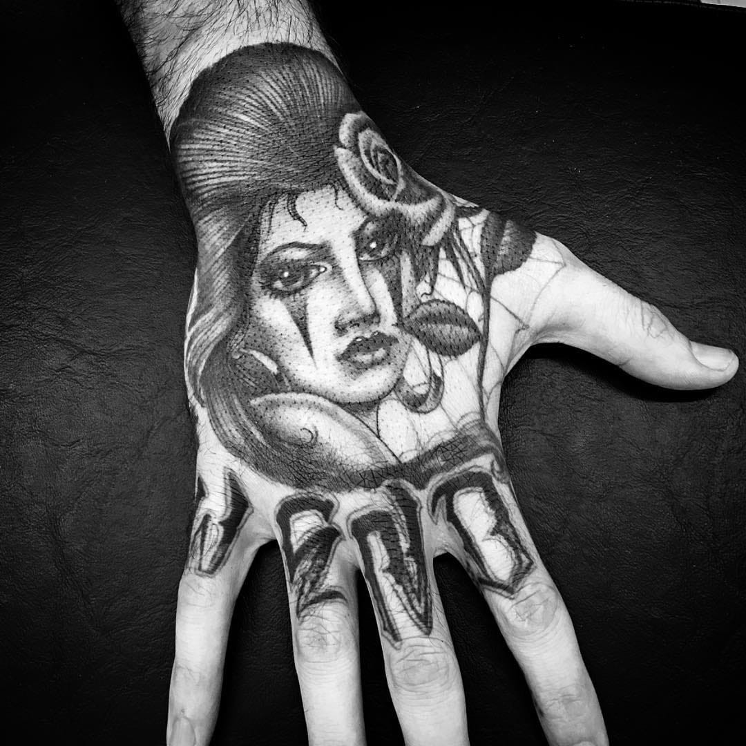 Tattoo by Chuco Moreno. Hand Chicano tattoo by Chuco Moreno featuring a payasa, lady head, rose, and lettering in black and grey, showcasing the intricate detail and placement of Chicano hand tattoos.