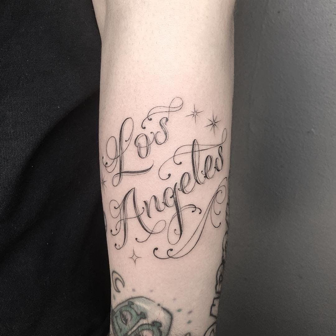 Tattoo by Em Scott. Script Chicano tattoo by Em Scott with Los Angeles lettering and stars in black and grey, showcasing the signature script and urban themes in Em Scott's Chicano tattoo style.