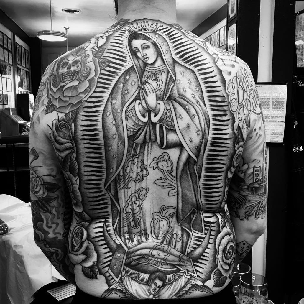 Tattoo by Chuco Moreno. Classic Chicano tattoo by Chuco Moreno featuring the Virgin Mary, roses, and a sugar skull in black and grey, showcasing the religious and cultural iconography prevalent in Chicano tattoo art.