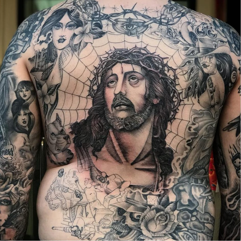 Tattoo by Big Steve. Complex Chicano sleeve tattoo by Big Steve featuring Jesus, a gun, payasa, roses, skull, cholo, barbed wire, spiderweb, pitbull, and crown of thorns in black and grey, demonstrating the detailed and symbolic nature of Chicano sleeve tattoos.