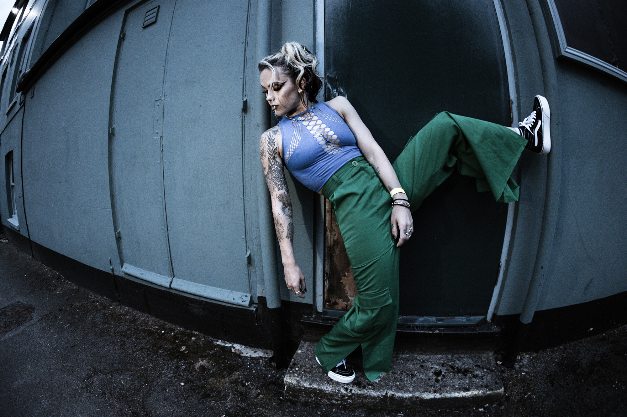 Wren in a black top, standing in front of a brick wall, looking to the side with tattoos visible on her arms