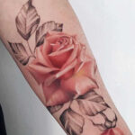 Realistic red rose tattoo with detailed shading on inner forearm, featuring black and grey leaves.