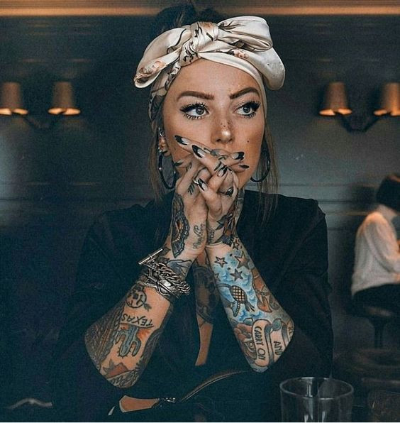 Tattooed woman with headscarf and clasped hands sitting in a dimly lit room.