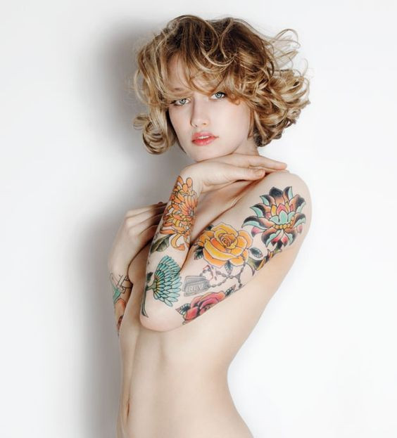 Woman with blonde curly hair showcasing colorful flower tattoos on her arms against a plain white background.