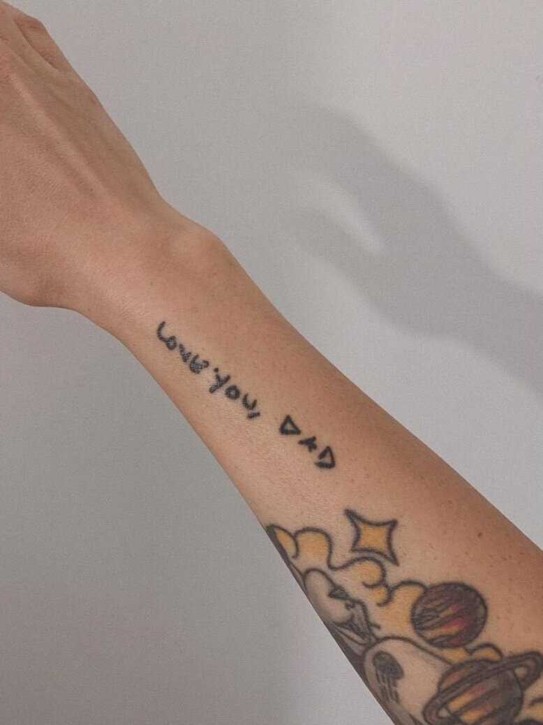 Close-up of Casha's forearm tattoo, featuring the handwritten phrase &quot;I love you, dad&quot; in black ink, a memorial to her father.