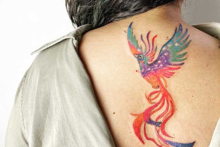 A vividly colored phoenix tattoo on a woman's back, showcasing an artistic blend of orange, red, yellow, and purple hues to express rebirth and personal artistic style through body art.