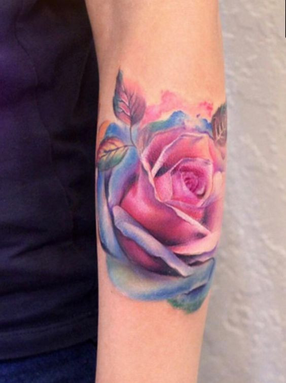 Vibrant watercolor rose tattoo on an arm, showcasing detailed petals in shades of pink, purple, and blue.