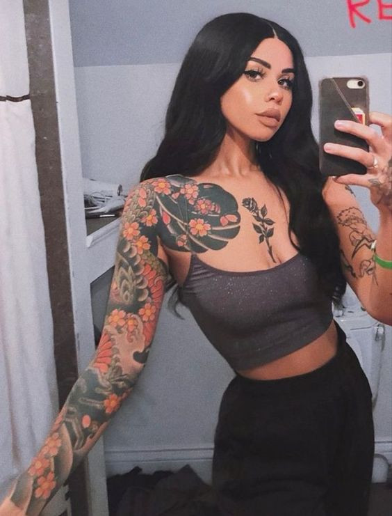 Woman with long black hair taking a mirror selfie, showcasing her colorful floral tattoos on her arm and shoulder.