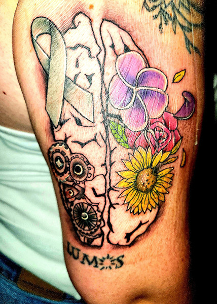 A strength tattoo showing a brain with mechanical gears on one side and flowers on the other, adorned with a gray ribbon for alt text &quot;Intricate strength tattoo of a brain divided by gears and flowers, symbolizing logic, creativity, and brain tumor survivorship&quot;.