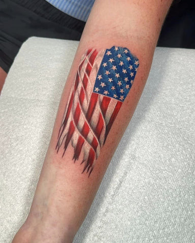 Vibrant American flag tattoo on a man's outer forearm, waving dynamically with detailed stars and stripes.