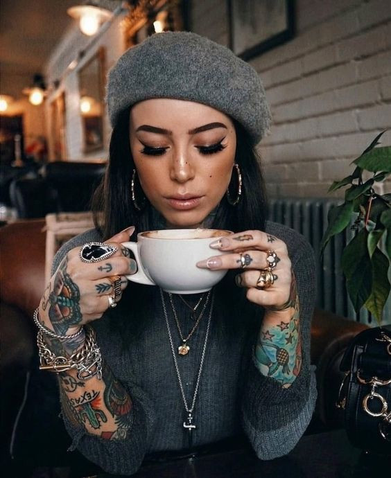 Woman with tattoos and jewelry enjoying a cup of coffee in a cozy cafe, wearing a grey beret and stylish outfit.