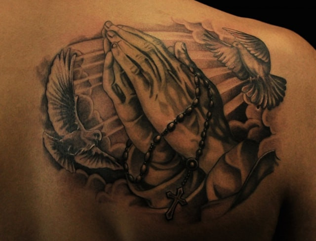 A back shoulder tattoo in black and grey ink, featuring praying hands clasped together with doves flying around them in a detailed and shaded style.