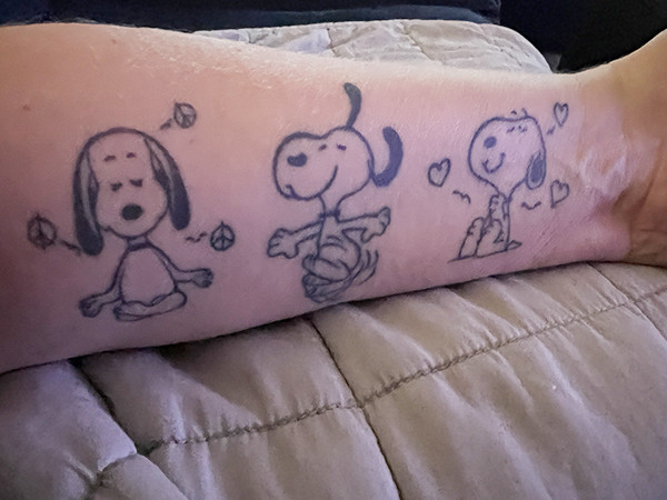 Three Snoopy tattoos on Cathy O.'s arm: Meditative Snoopy, Dancing Snoopy, and Radiating Love Snoopy, representing life lessons learned from her journey.