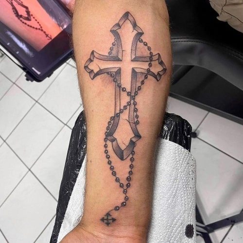 Two cross tattoos on men, one on the forearm with a detailed Celtic cross design and another on the chest with a large ornate cross with wings.