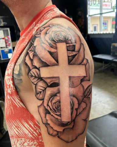 Detailed black and gray cross tattoo on a man's shoulder, incorporating shading and realism.