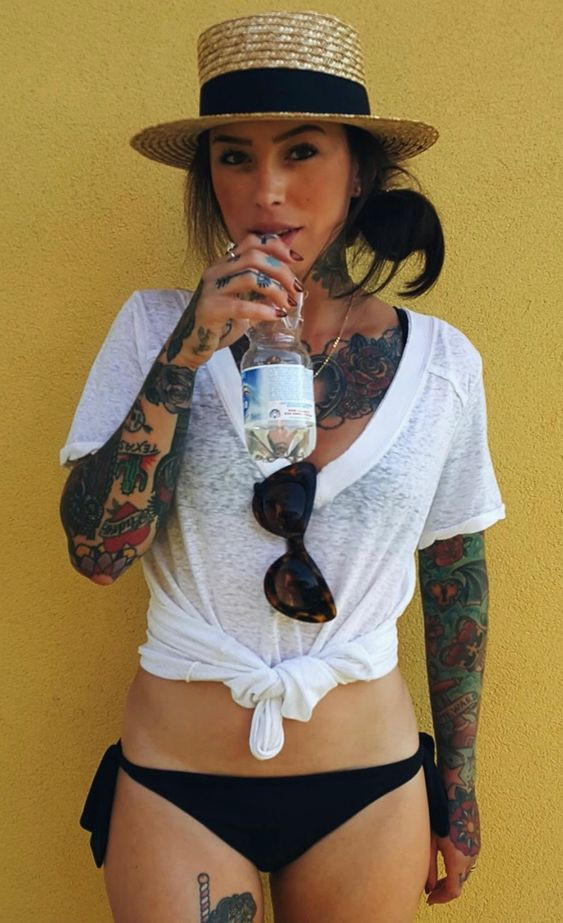 Woman with tattoos in a white shirt, black bikini, and straw hat drinking water against a yellow background.