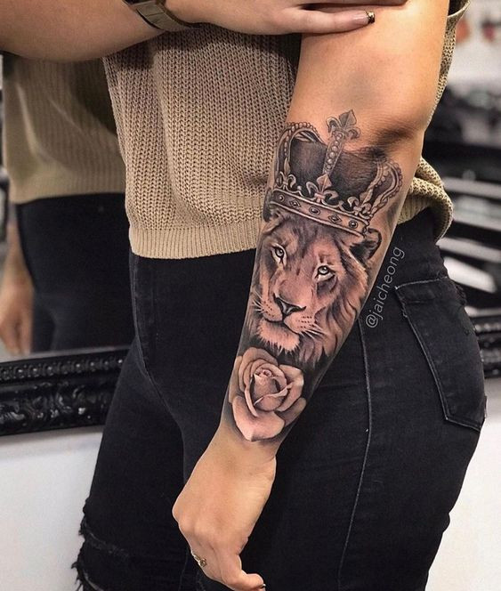 Arm tattoo of a crowned lion and rose on a person wearing a beige top and black jeans.