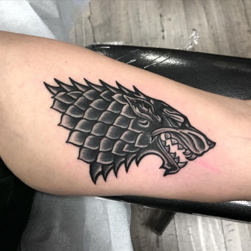 A dire wolf tattoo on a man's arm, depicting a larger, more imposing wolf with a prehistoric feel, symbolizing ancient power and loyalty inspired by mythology and popular culture.