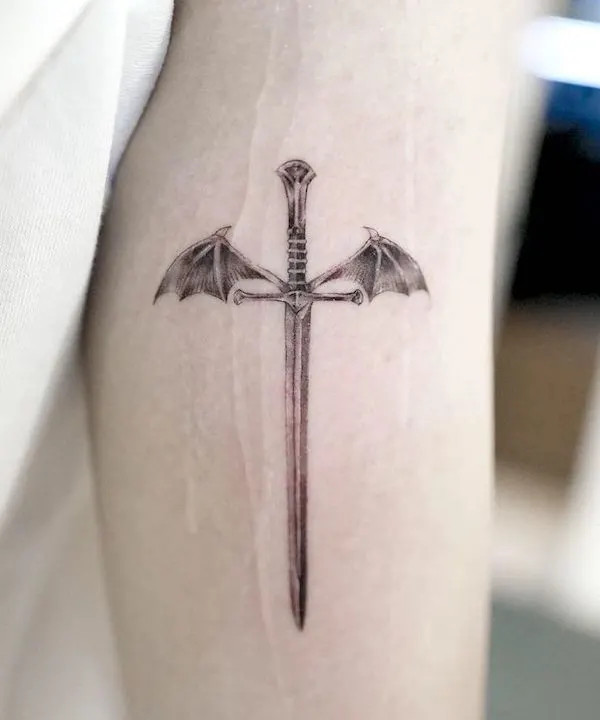 alt text: Small dragon wing and sword tattoo on arm in black ink, symbolizing strength, confidence, and fighting spirit