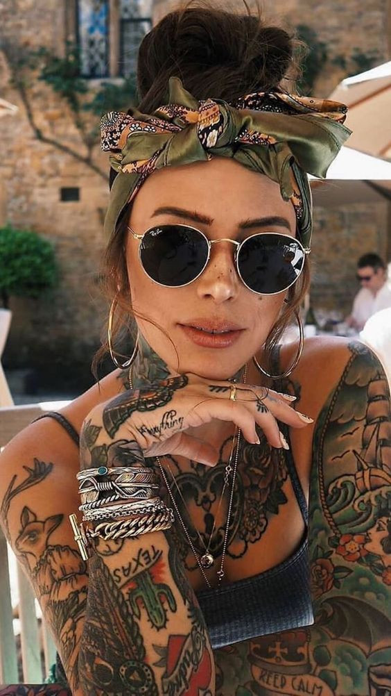 Tattooed woman with sunglasses, headscarf, and bracelets posing outdoors in sunny weather.