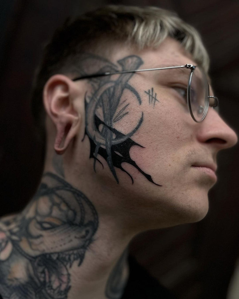 Cool face tattoo featuring a delicate feather design extending from the hairline to the cheek in black ink.