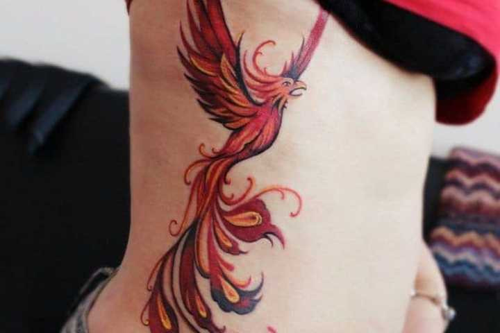 A dynamic rising phoenix tattoo on a woman's side, vividly colored in orange, red, and white to capture the energy of rebirth, transformation, and triumph over adversity through body art.