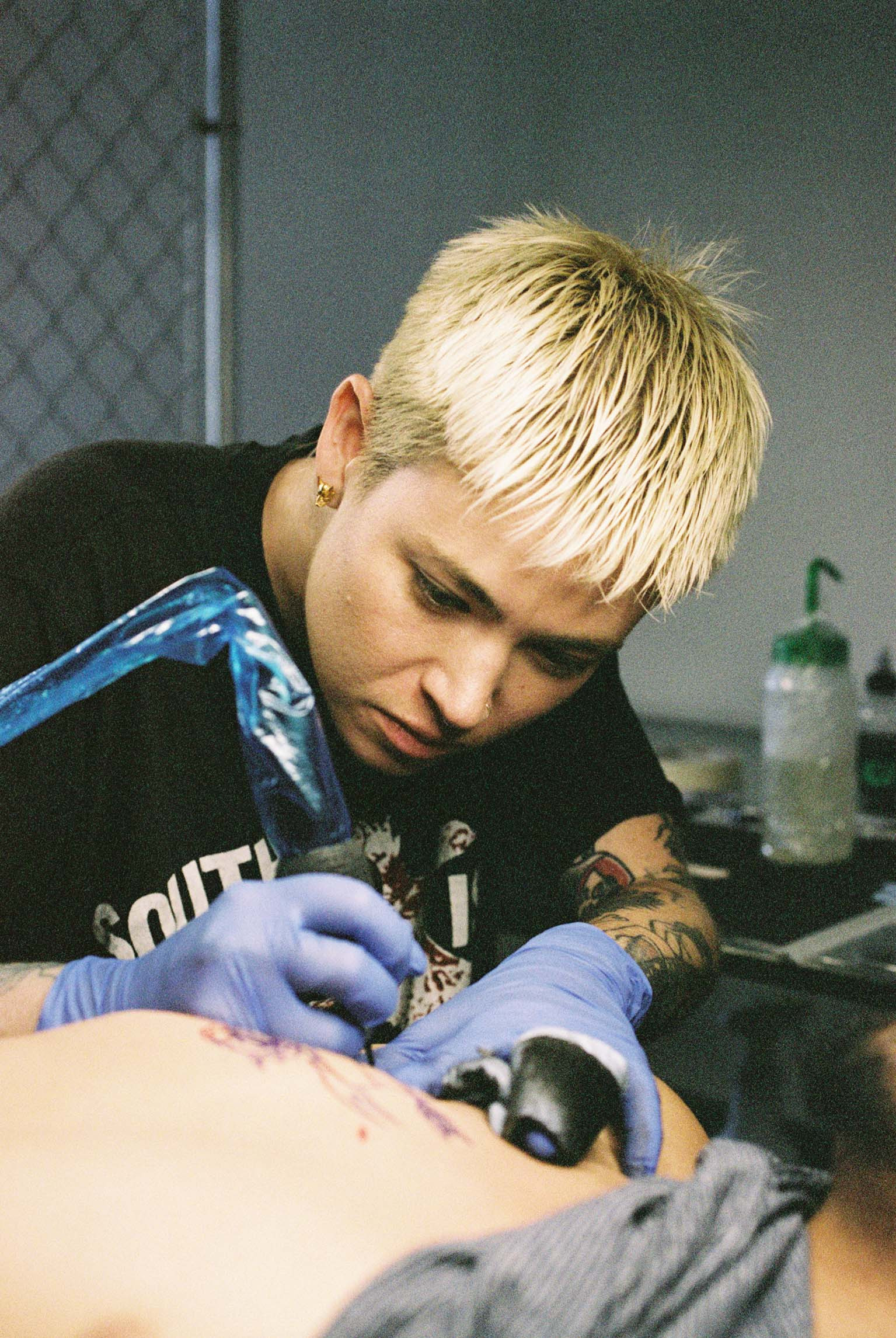 Close-up shot of Bobby Coyote's tattooed arm and hand, holding a tattoo machine, focused on tattooing skin