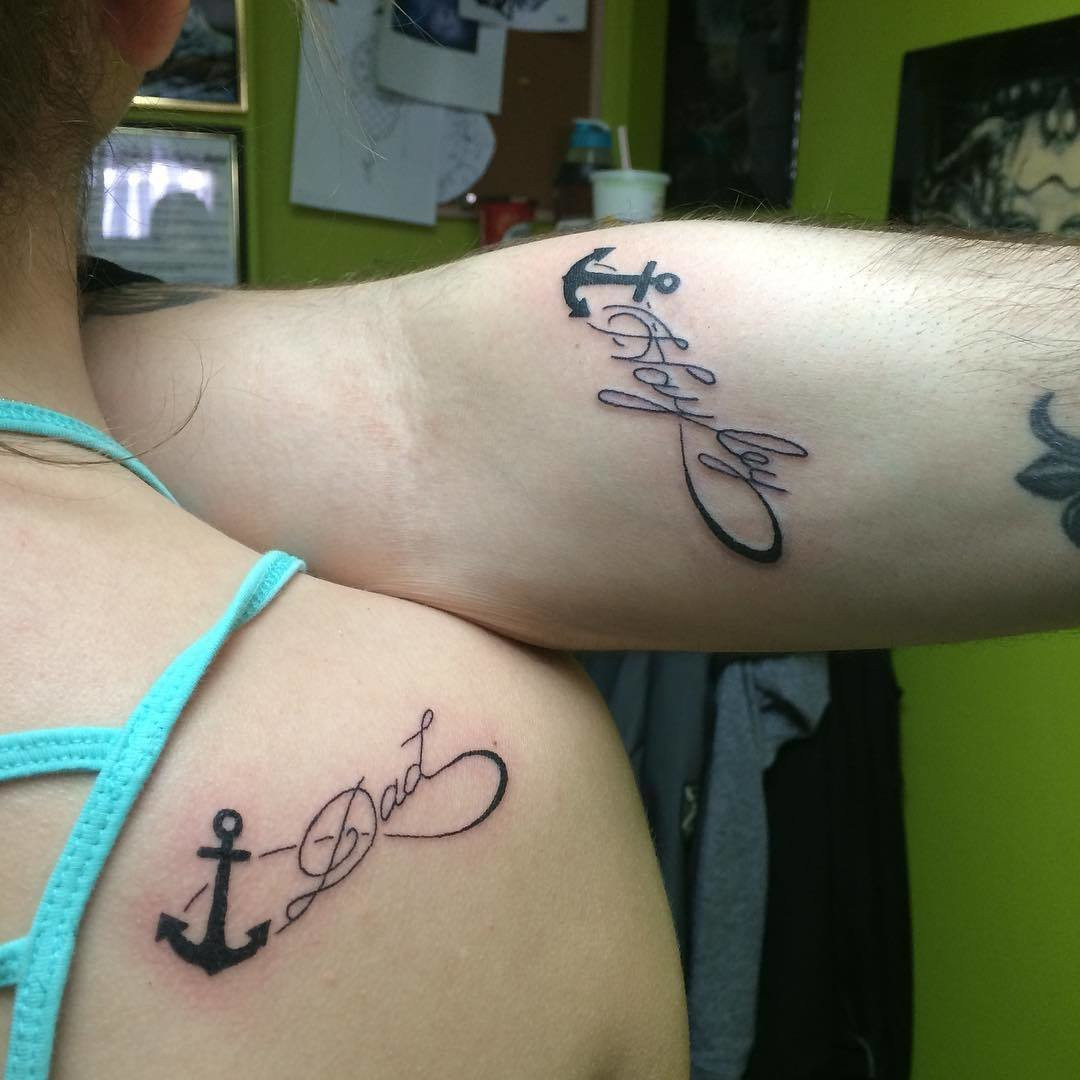 Father and daughter showcasing wrist tattoos of infinity symbols incorporating the word &quot;dad&quot; and a daughter's name, along with an anchor symbol, representing everlasting love and stability.