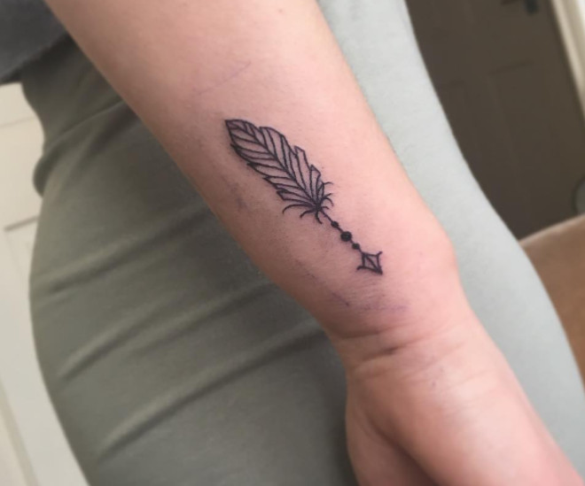 A small black ink feathered arrow tattoo positioned on the outer wrist.