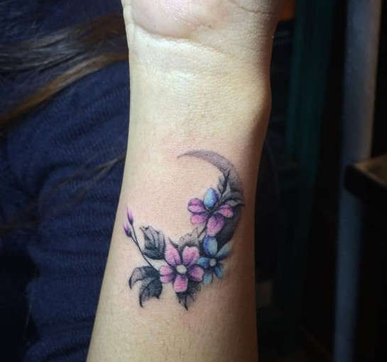 A small flower and moon tattoo in grayscale with pink and blue accents on a wrist.