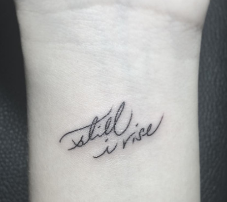 A wrist tattoo with the quote &quot;Still I Rise&quot; in a delicate font.