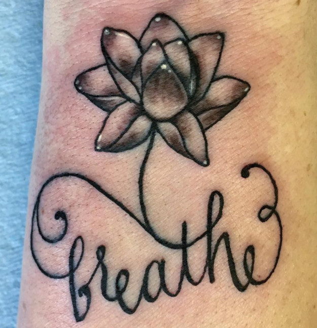 A lotus flower and the word &quot;breathe&quot; tattoo in grayscale on a wrist.