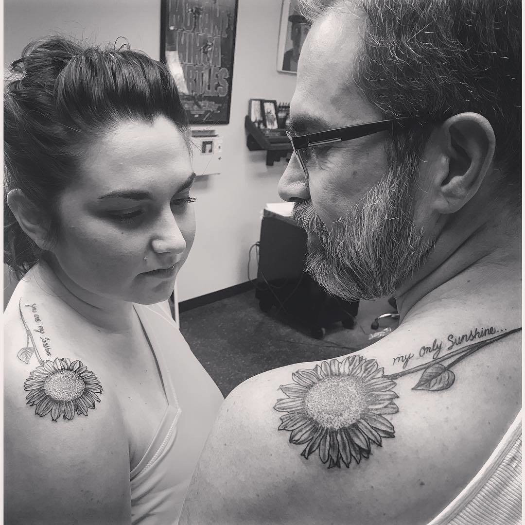 Father and daughter displaying shoulder tattoos of sunflower designs, symbolizing adoration and loyalty, enhanced by &quot;You Are My Sunshine&quot; lyrics.