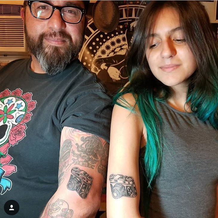 Father and daughter displaying forearm tattoos of camera designs, symbolizing their shared passion for photography and capturing memories together.