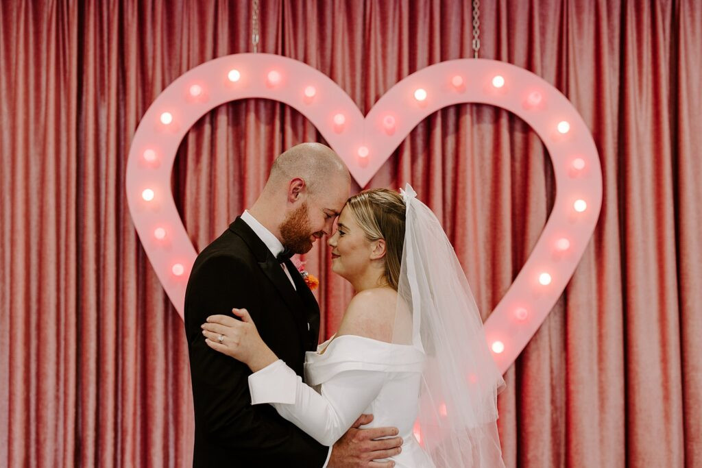 bride and groom touch foreheads in front of pink heart before going to Downtown Tattoo shop