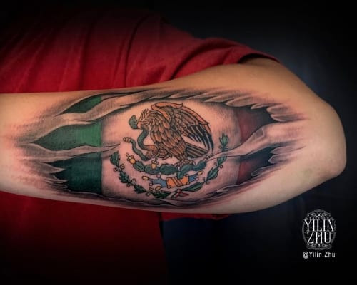 Two mexican tattoos on men, one on the forearm with a sugar skull and roses and another on the arm with a Mexican flag and eagle.