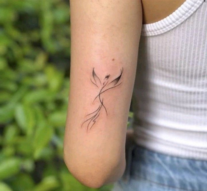 Minimalist phoenix tattoo on upper arm, worn by a person in a white tank top, with a leafy green background.