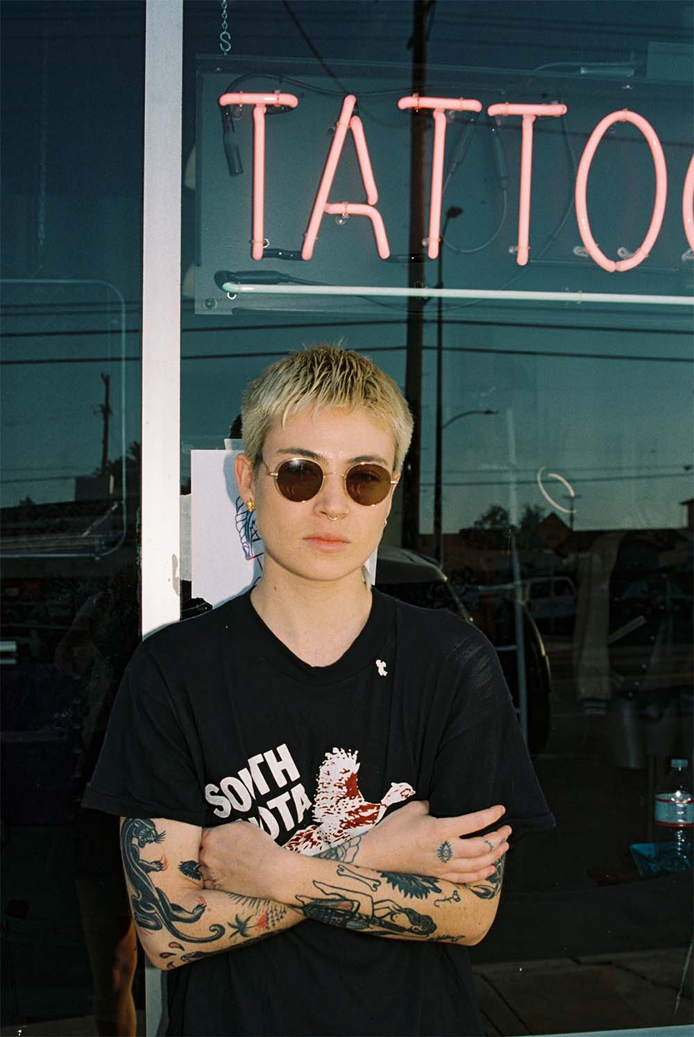 Bobby Coyote wearing sunglasses and a denim jacket, holding a cigarette and smiling, showcasing a relaxed and cool style