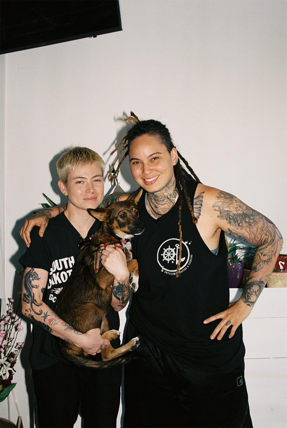 Bobby Coyote with short hair and a black shirt, looking directly at the camera with a serious expression, tattoos visible on their neck and arms