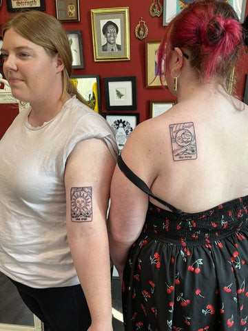 Two friends displaying their matching tarot card tattoos, one with a sun design and the other with a moon, inked by Sarah Louise to represent balance in friendship.