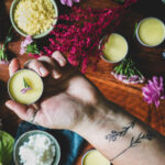 A hand hold a tin of homemade tattoo balm above a table that is strewn with ingredients and flowers