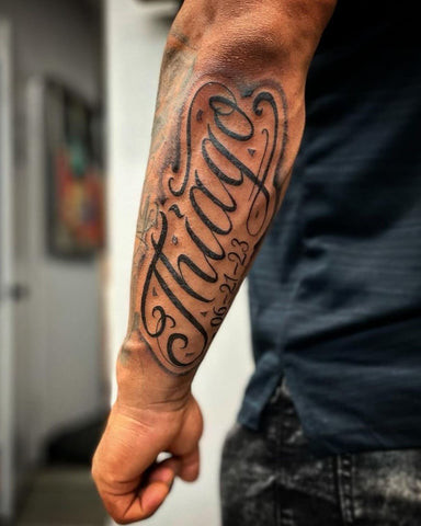 Elegant script name tattoo on a man's outer forearm, featuring flowing cursive lettering in black ink.