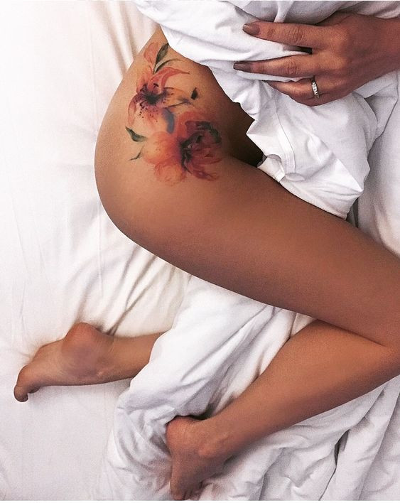 Floral hip tattoo on woman lying in bed with white sheets, showcasing colorful design.