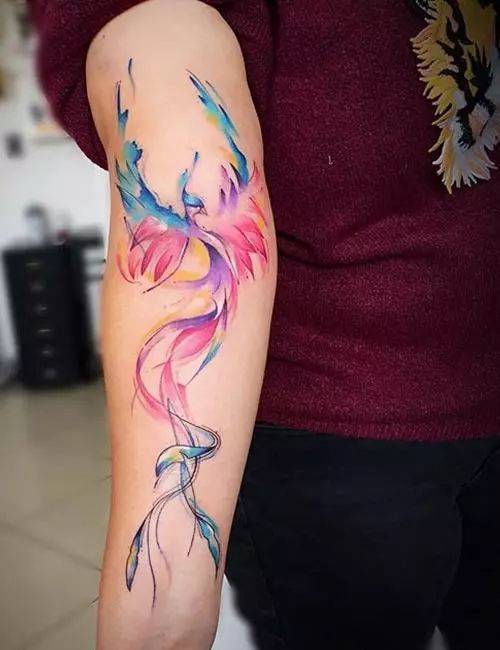 Colorful watercolor phoenix tattoo on forearm, blending shades of purple, blue, and pink against a maroon shirt background.